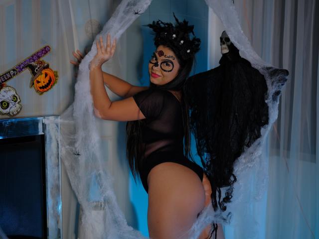 Beckyte con máscara negra, cabello largo y top negro transparente, de espaldas a una cortina, mostrando su trasero, usando alas negras, en una foto con temática de Halloween, con las manos en la cortina..