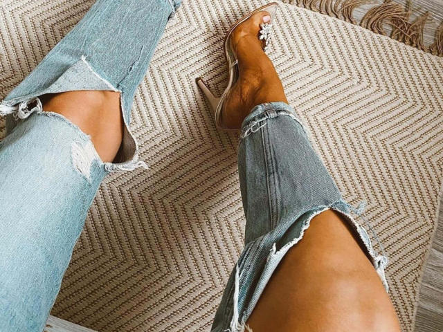Le jean bleu déchiré de ShanonHorny présente des ourlets effilochés, et ses chaussures marron à talons hauts et ouvertes sont ornées de clous argentés, le tout sur un tapis beige texturé..
