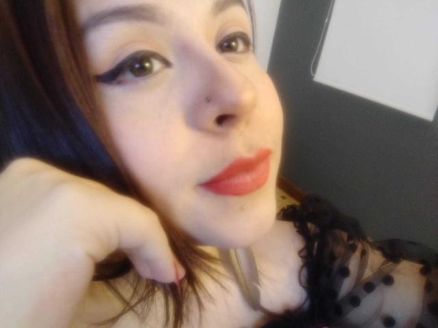 Le visage de Katie Woolf, avec du rouge à lèvres rouge, de l'eye-liner noir et un piercing au nez, tenant ses cheveux bruns, portant un haut noir transparent à pois, laissant entrevoir ses seins..