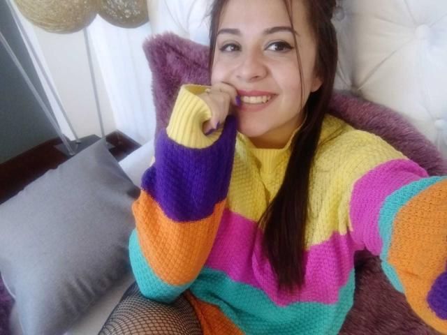 Katie Woolf, con coletas marrones, sonriendo, vistiendo un suéter tejido a rayas de colores, esmalte de uñas morado y medias de red, tocándose la barbilla con una expresión linda y juguetona..