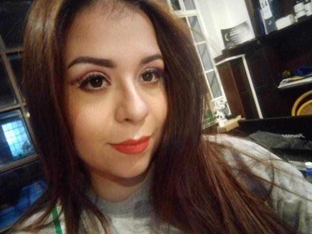 Katie Woolf ha gli occhi marroni, i capelli lunghi castani, il rossetto rosso e un incarnato chiaro. Indossa una maglietta grigia e guarda la fotocamera con un leggero sorriso..