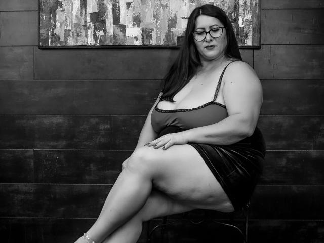 SoniaLaBelle, plus-size with big tits, black dress, and glasses, sits crossed-legged on a bench, black wall and abstract painting behind, black and white photo.