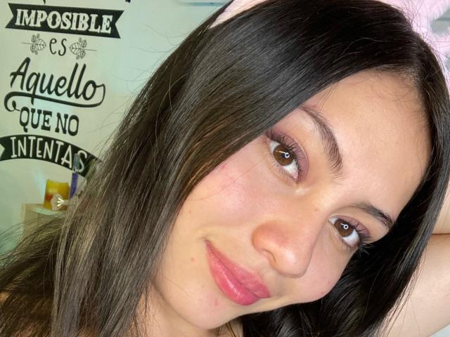 AlexaCoeur has brown eyes, pink lips, and black straight hair, smiling with a slight tilt. Her skin is fair with a small red blemish on her right cheek.