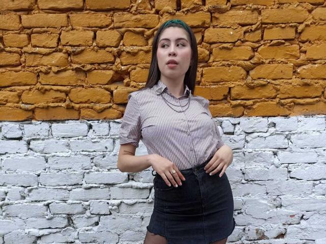 IsaTimeSweet, with green headband, striped shirt, and black skirt, standing against a yellow and white brick wall, hands on hips, looking up with a confident expression.