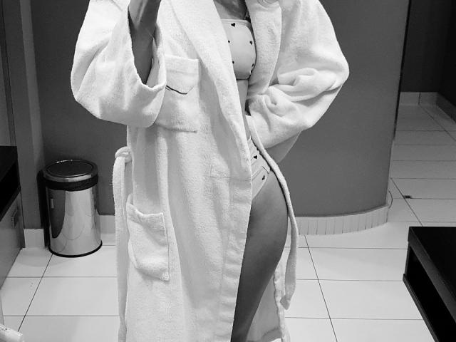 Fotografía en blanco y negro de LenaaDoll vestida con una bata blanca esponjosa, que revela un sujetador y bragas con lunares, con una mano en la cadera, mostrando un trasero pequeño y lindo..