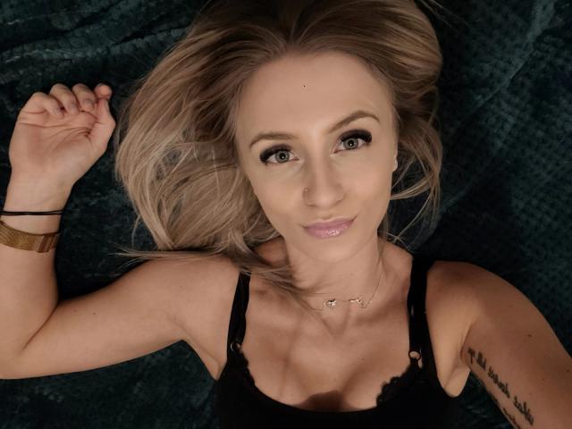 LenaaDoll, com cabelo loiro, olhos azuis e um piercing no nariz, deitada em um cobertor verde escuro, vestindo uma blusa preta de renda e um bracelete dourado, sorrindo para a câmera..