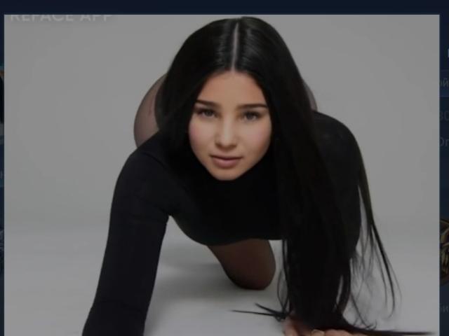 LuxeDerta is on all fours, wearing a black long-sleeve top, with long black hair, looking forward with a slight smile, showing her ass and face.