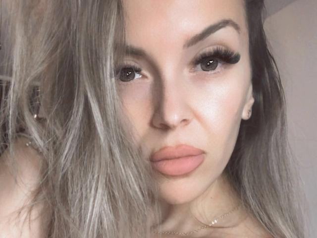 SophieDeee's face with full lips, grayish-blonde hair, and thick, long eyelashes. She's wearing a gold necklace and has a serious, slightly pouty expression. Her tits are not visible.