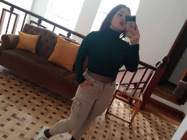Katie Woolf, con i suoi lunghi capelli castani, il maglione verde a collo alto e i pantaloni beige, mostra il suo sedere in un selfie allo specchio, tenendo un telefono, in piedi su un pavimento a motivi..