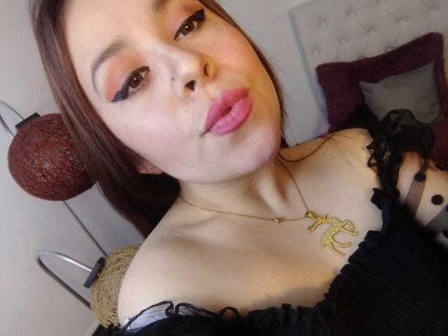 Katie Woolf, con le labbra piene, gli occhi marroni e l'eyeliner nero, indossa una maglietta nera con una spalla scoperta, una collana dorata con un ciondolo a forma di chiave e un piccolo piercing al naso..