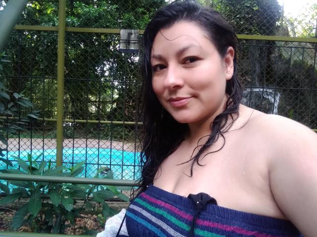 SakotoDika has wet black hair, a slight smile, and visible small tits in a strapless blue and purple striped top, standing by a green fence and a blue pool.