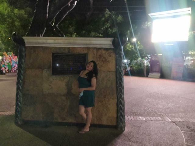 SakotoDika with dark hair, green dress, and sandals, standing in front of a stone wall at night, leaning with hands on wall, looking to the side, ass slightly visible.