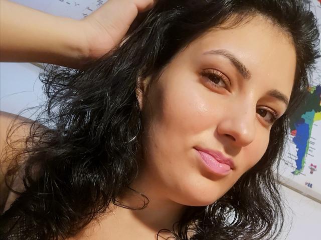 AddictedToUTS has wavy black hair, brown eyes, pink lips, and a light tan skin tone. She's touching her head with a hand, looking slightly to the side.