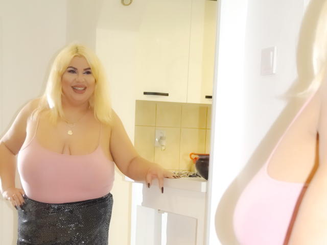 HornyBbw with blonde hair, pink tank top showing big boobs, black sequined skirt, smiling, leaning on white door, reflection in mirror. Blonde reflection in foreground.