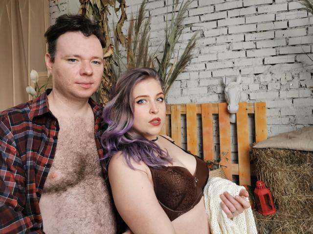 Genrikh Becca, com cabelo roxo, um sutiã de renda marrom e um suéter branco, está ao lado de um homem com peito peludo, vestindo uma camisa xadrez, e ambos estão olhando para a câmera..