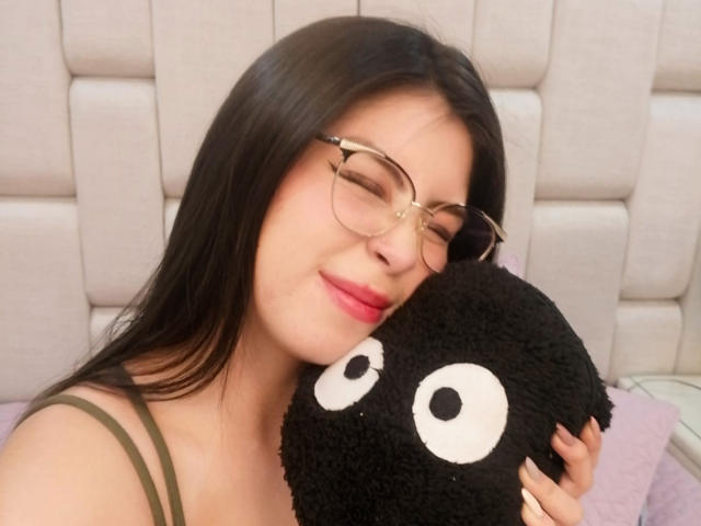 IsaTimeSweet, com cabelos pretos longos, óculos e batom rosa, segurando um travesseiro preto e fofo com olhos brancos, sorrindo suavemente, vestindo uma blusa verde com alças, parecendo adorável..