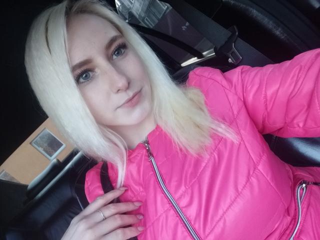 EllenSoNice, met blond haar, blauwe ogen en een roze, opvallende jas, laat haar kleine borsten en schattige gezicht zien in een auto. Ze houdt de jas vast met een ring..