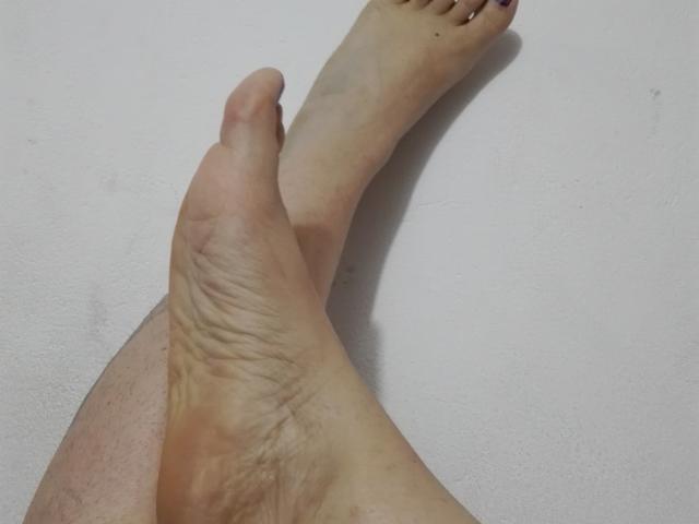 De voet van een LatinaHardGirl, met zichtbare rimpels, een licht vuile zool en paars geschilderde teennagels, steekt over een andere voet op een witte ondergrond..