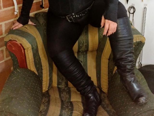 LatinoHardGirls in black leather pants and thigh-high boots, sitting on green and yellow striped chair, black belt with studs, arms resting on chair, brick wall background.