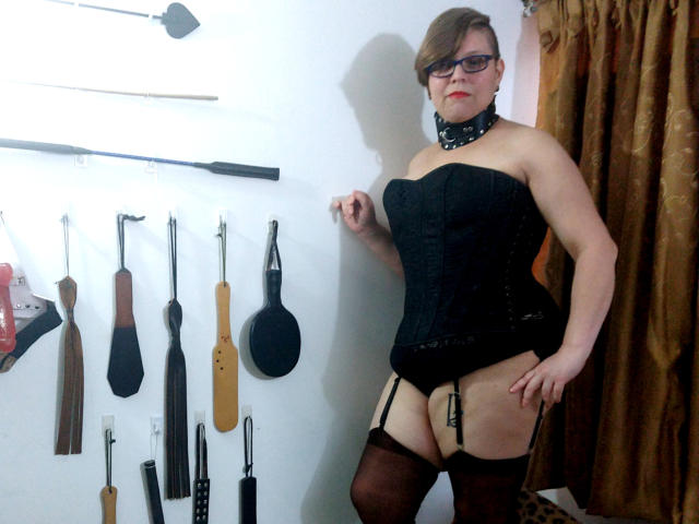 Mulheres latinas com cabelo castanho curto, óculos, corpete preto, cinta de suspensórios, meias e colar, em pé, mostrando os seios, brilhando com uma vibe confiante, sexy e dominante, em uma sala BDSM..