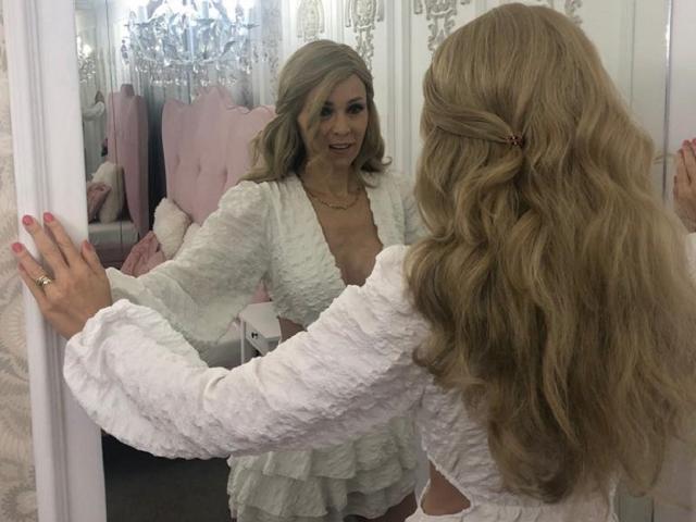 Kassandra Gray, met golvend blond haar, een witte, gestructureerde jurk en roze nagels, glimlacht naar haar spiegelbeeld, waarbij haar borsten en billen in de spiegel zichtbaar zijn..