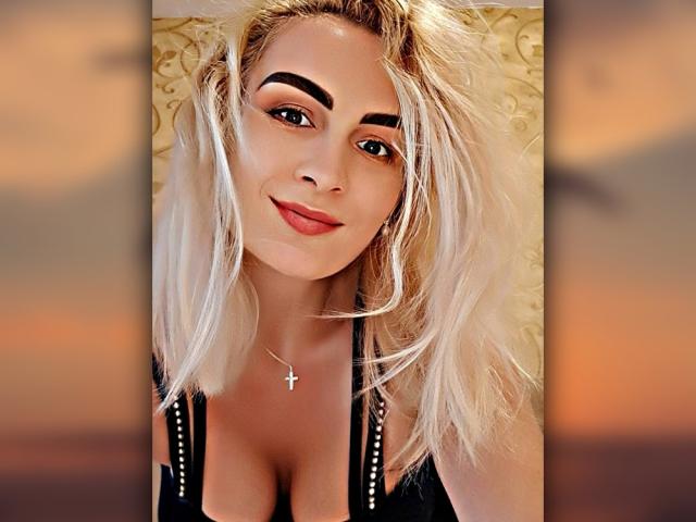 Sexy vrouw met blond haar, dikke wenkbrauwen, rode lippenstift, zwarte top, diep decolleté, kruisbedel, lachend, selfie, zonsondergang op de achtergrond, onscherpe randen, kijkt naar de camera..
