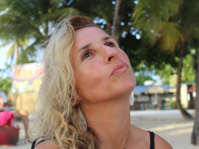 TitaLove a les cheveux blonds et ondulés, une peau claire, et elle fait une moue en regardant vers le haut. Elle porte des boucles d'oreilles noires et un haut noir..
