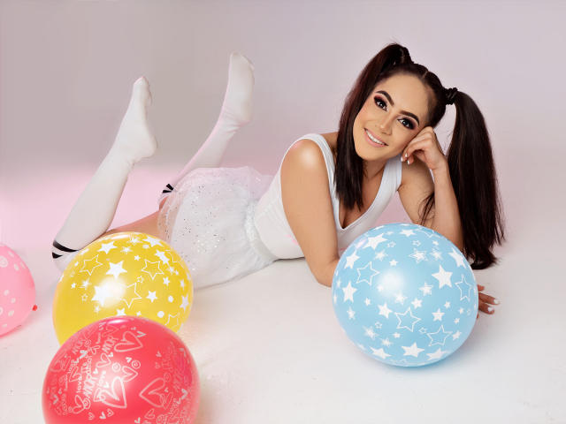 LunaRuiiz, com maria-chiquinhas, camiseta branca e meias brancas, está deitada de bruços, sorrindo, cercada por balões coloridos com estampas de estrelas, amarelos, rosa, vermelhos e azuis..