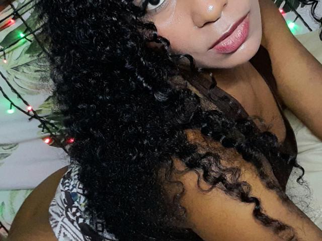 HottBianca69, con cabello negro rizado, un piercing en la nariz y lápiz labial rosa, acostada en una cama, vistiendo ropa interior estampada, mirando a la cámara con una ligera sonrisa..