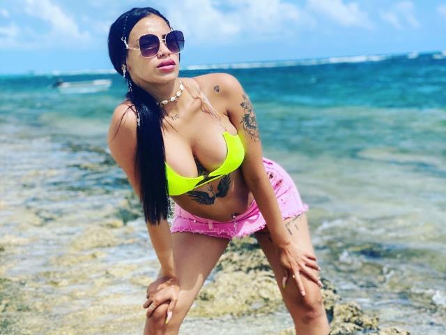 OnniOconor with large tits in a neon yellow bikini top, pink shorts, tattoos, and sunglasses, bending forward on a rocky beach, ocean in background.