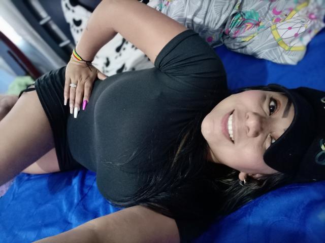 AvaSexySeins deitada em uma cama azul, vestindo uma camisa preta e shorts, um lenço preto na cabeça, sorrindo, mostrando seios grandes, unhas cor-de-rosa, uma pulseira amarela, um anel e alargadores de orelha..