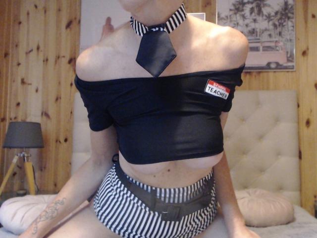FrenchPlumeX with bare tits, black off-shoulder top, striped skirt, and black-and-white tie, sitting on a bed, wooden walls, tattoo on left arm.