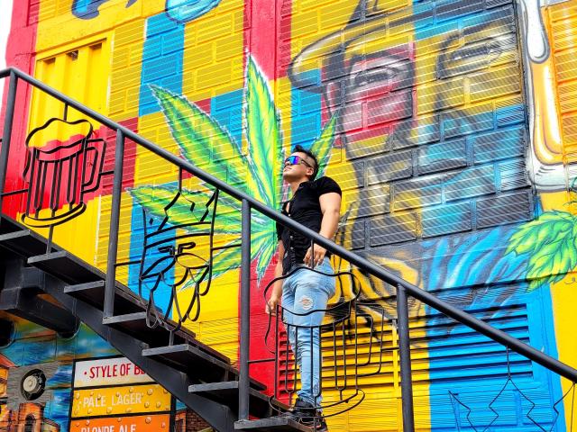 AngelLin è in piedi su delle scale di metallo, indossa una maglietta nera e jeans strappati, con occhiali da sole verdi, di fronte a un muro colorato pieno di graffiti. Il suo sedere è leggermente visibile, mentre si volta a guardare..