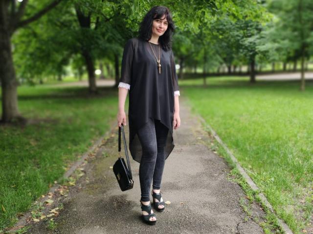 Gusum with black shirt, gray pants, and green shoes, holding black bag, wearing necklace, standing on path, green trees in background, looking down, casual outfit, medium-length black hair.