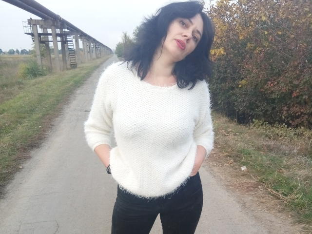 Gusum has black wavy hair, red lipstick, and a fluffy white sweater, hands in black pants, standing on a paved path, looking slightly upward with a subtle smile.