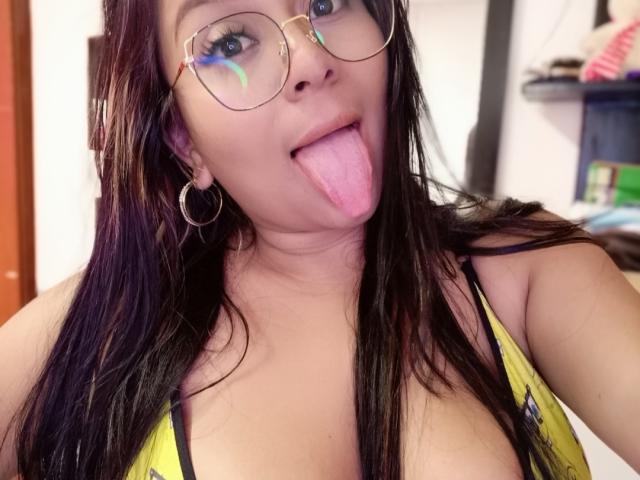 AvaSexySeins with long black hair, glasses, hoop earrings, and yellow top, sticking out her pink tongue. Her boobs are visible, and she's looking at the camera.