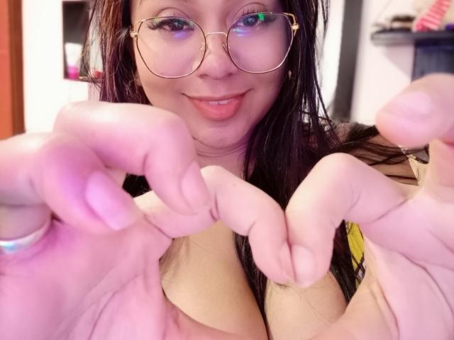 AvaSexySeins mit Brille, lächelnd, zeigt ihre Hände, die ein Herz formen, rosafarbener Ring am Finger, langes braunes Haar, helle Haut, rosafarbener Lippenstift und deutlich sichtbare große Brüste..