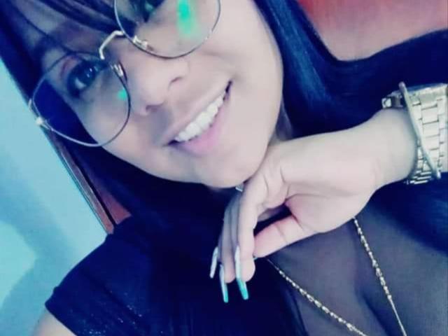 AvaSexySeins com óculos, sorrindo, cabelo preto, unhas azuis, relógio dourado, blusa preta, peito visível, apontando para o queixo, usando um colar dourado, parecendo fofa..