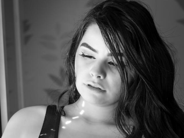 Black-and-white photo of BrithneyLee with long, dark hair, closed eyes, and winged eyeliner, wearing a strap dress, looking slightly down, light reflecting on her chin.