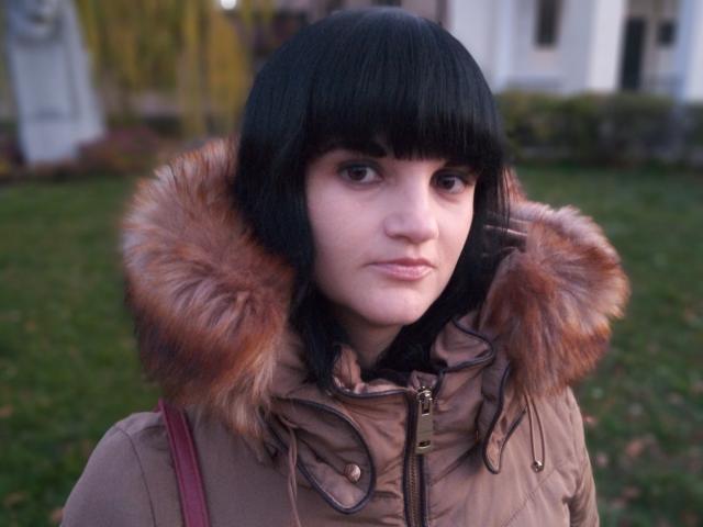 XLylyusX, con cabello negro corto, vestida con un abrigo marrón abultado con capucha adornada con piel, está de pie en un césped, tiene piel pálida y una expresión neutra..