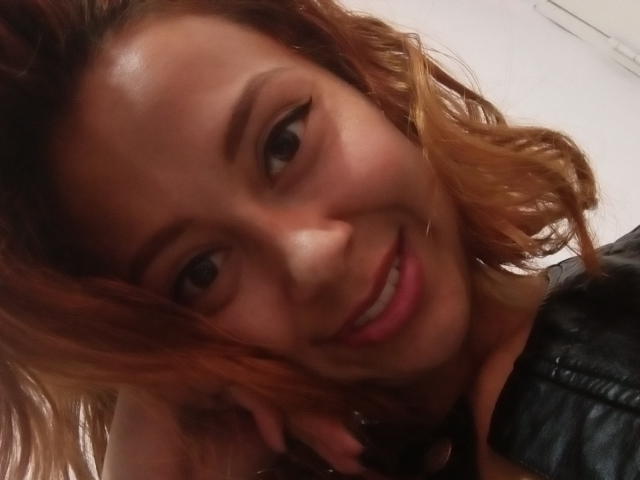 Eliza Jones, con cabello castaño ondulado, sonriendo, vistiendo una chaqueta de cuero negra, mostrando sus labios rosados y sus ojos marrones, en un selfie ligeramente borroso..