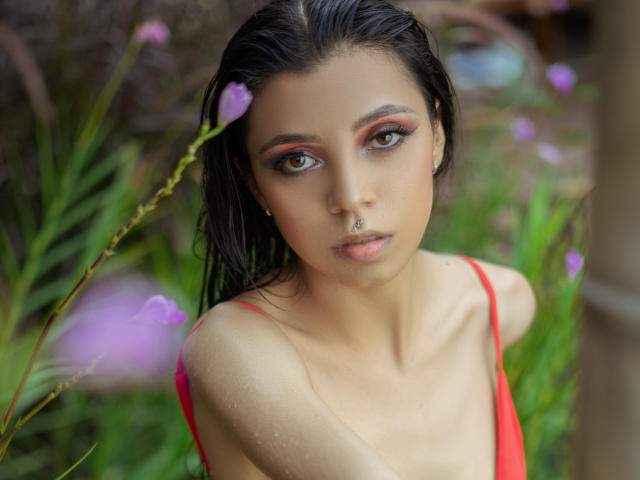 VannesaFlory, con i capelli neri bagnati, il trucco rosa sugli occhi e un piercing al naso, indossa una maglietta rossa, guarda verso la fotocamera, con dei fiori viola sullo sfondo..