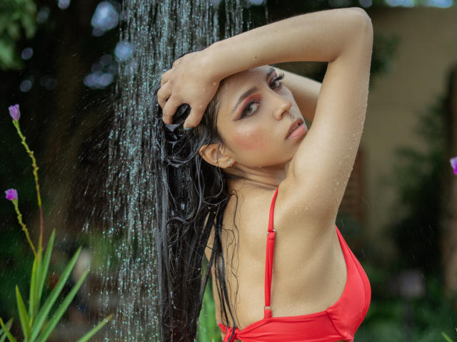 VannesaFlory, com cabelo preto molhado, braços acima da cabeça, maiô vermelho, gotas de água, olhando para trás, folhas verdes, flor roxa, expressão sensual, seios médios, atraente, ambiente externo..