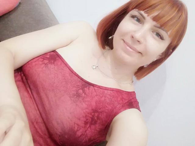 LaylaHottyX mit rotem Bob, trägt ein rotes Tie-Dye-Top, zeigt kleine Brüste, lächelt, hat helle Haut, eine silberne Halskette, Ohrringe und dezentes Make-up, auf einem Selfie..