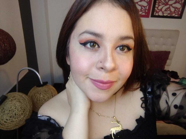Katie Woolf ha la pelle chiara, gli occhi marroni e il rossetto rosa, indossa pizzo nero, una collana d'oro con una cerva, e ha una mano sul viso, sorridendo..