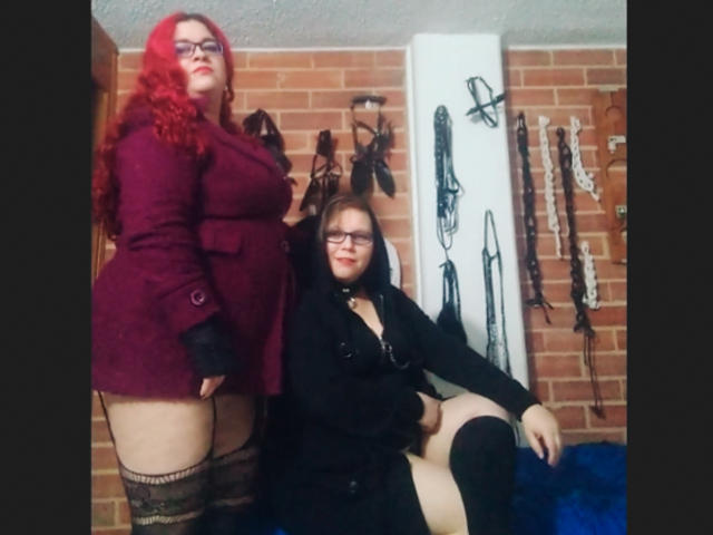 Duas latinas com cabelo ruivo e preto, óculos e roupas góticas. Uma está em pé, exibindo suas nádegas, em um casaco bordô. A outra está sentada, exibindo seus seios, em um vestido preto..