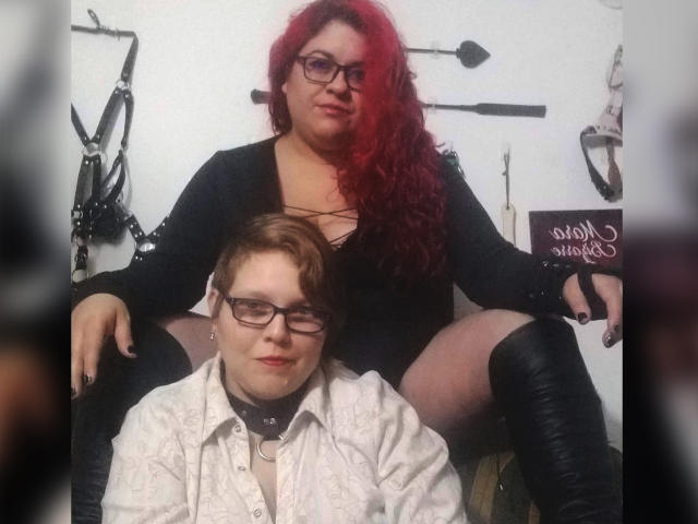 LatinoHardGirls mit roten Locken, schwarzem Kleid und Brille sitzen auf einem Stuhl, die Beine gespreizt, und zeigen ihre Vulva. Eine LatinoHardGirl mit Brille, weißem Hemd und Kragen sitzt davor..