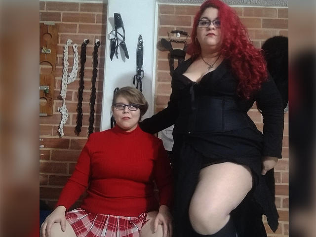 Duas latinas, uma com cabelo ruivo encaracolado, vestido preto e coxa à mostra, está em pé sobre a outra, que tem cabelo castanho curto, óculos, gola alta vermelha e saia xadrez..
