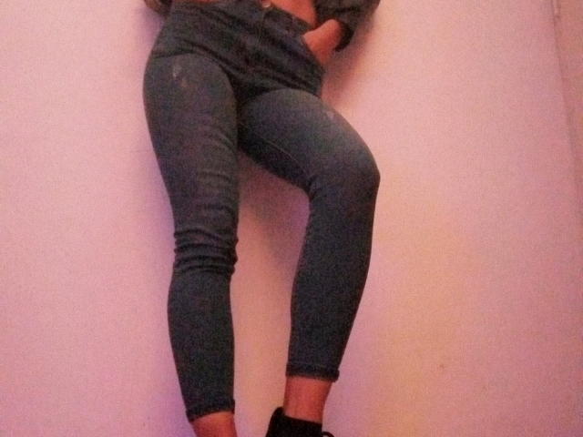 Eliza Jones está encostada em uma parede, vestindo jeans azuis justos e um moletom cinza, com a mão no bolso, pernas levemente cruzadas e meias pretas..