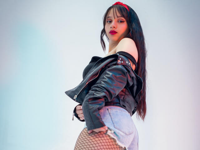 KloeThompson with long black hair, red headband, black leather jacket, red lipstick, fishnet stockings, and denim shorts, standing with a confident, slightly provocative pose.